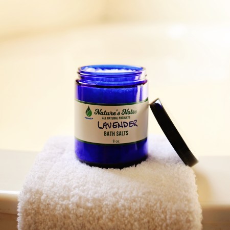 A photo of a cobalt blue jar with lavender bath salts sitting on the edge of a bathtub on top of a towel.